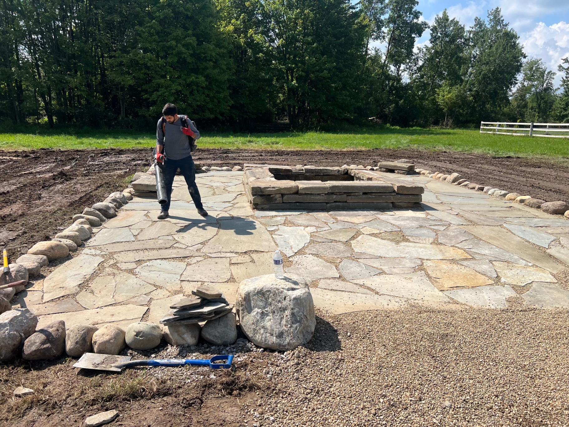 A natural flagstone patio with a stacked-stone fire pit and rounded boulder edging, with final cleanup underway.