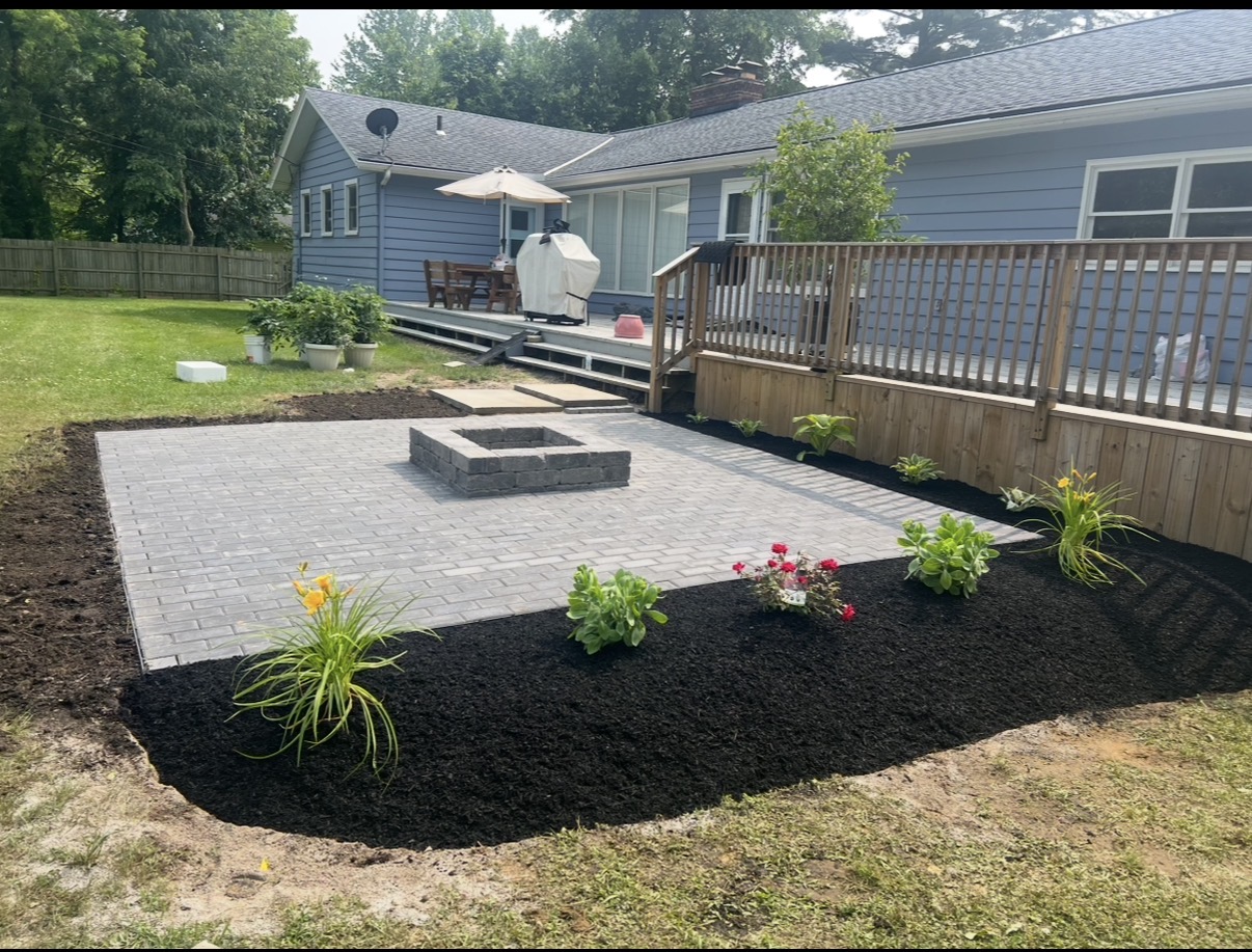 A gray paver patio with a square fire pit, bordered by fresh black mulch and new plantings.